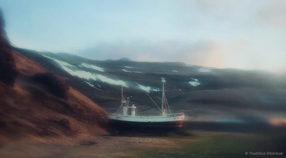 WhiteWall Magazine An old fishing boat stands on a wooded shore, surrounded by mountains.