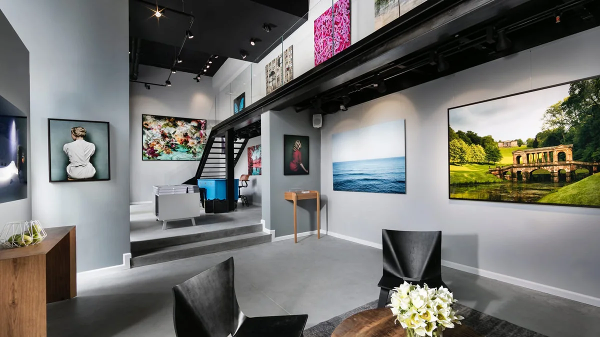 Business Customers Contact Form Interior view of a gallery with several works of art on the walls, including photographs and paintings.