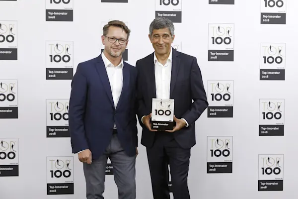 WhiteWall - Top 100 Innovation Award Two men in suits hold a Top 100 Innovation Award trophy in front of a sponsor wall.