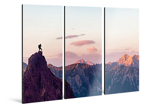 PDP | Multi Panel Wall Art | Images Text Section > Alu Dibond | Media #1 A man is standing on a rocky outcrop in front of a mountain panorama. The whole picture is divided into three parts hanging horizontally on the wall.